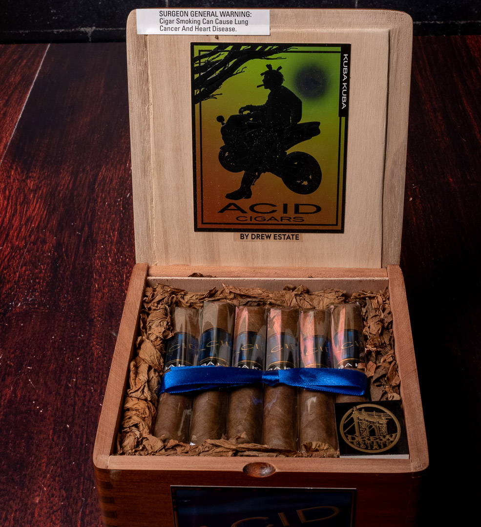 An open box of ACID Kuba Kuba cigars by Drew Estate, tied with a blue ribbon and nested in dried tobacco leaf