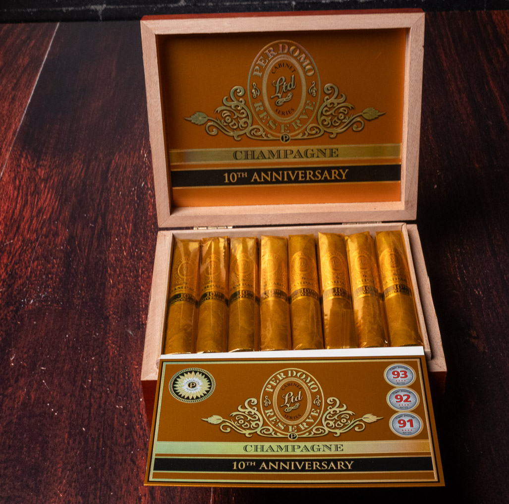 An open box of Perdomo 10th Anniversary Champagne Robusto cigars in gold foil sleeves on a dark wood table