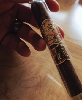 A hand holding a dark-wrapped cigar with an ornate red and gold band, still in its cellophane sleeve, over a wooden table