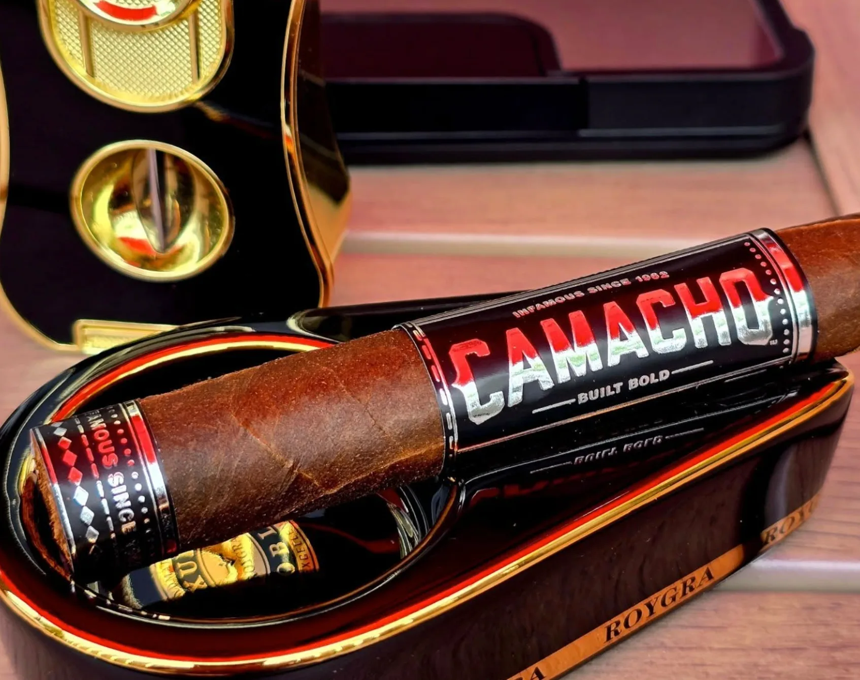 A Camacho cigar resting in a black and gold ashtray beside a cigar cutter
