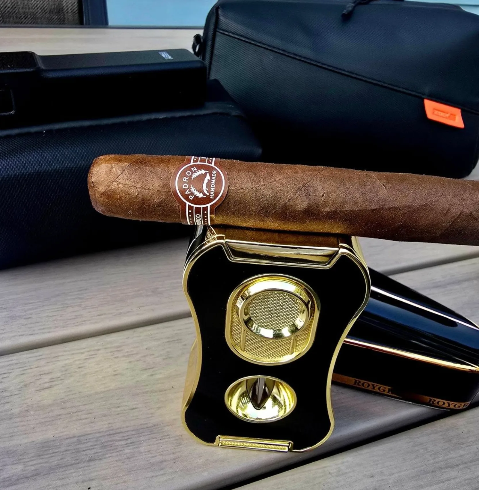 A Padrón cigar balanced on a black and gold cigar cutter on a wooden deck