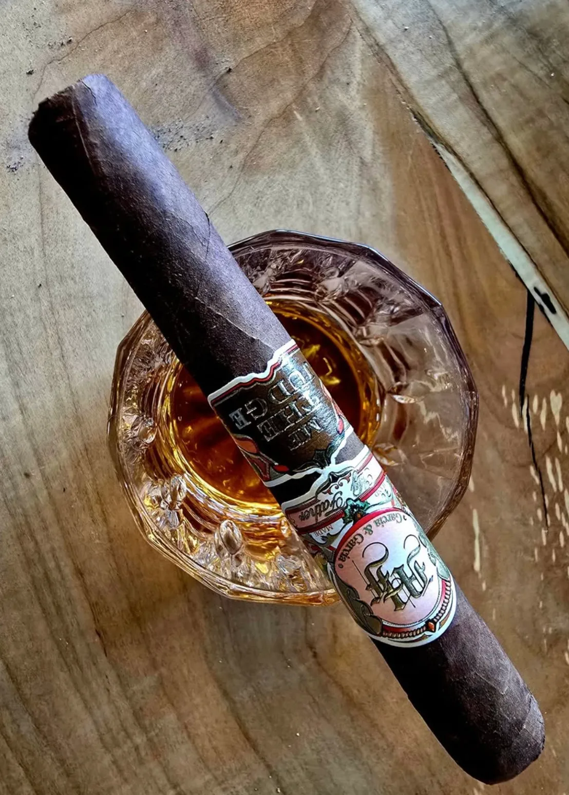 A dark-wrapper My Father cigar resting across a glass on a wooden table